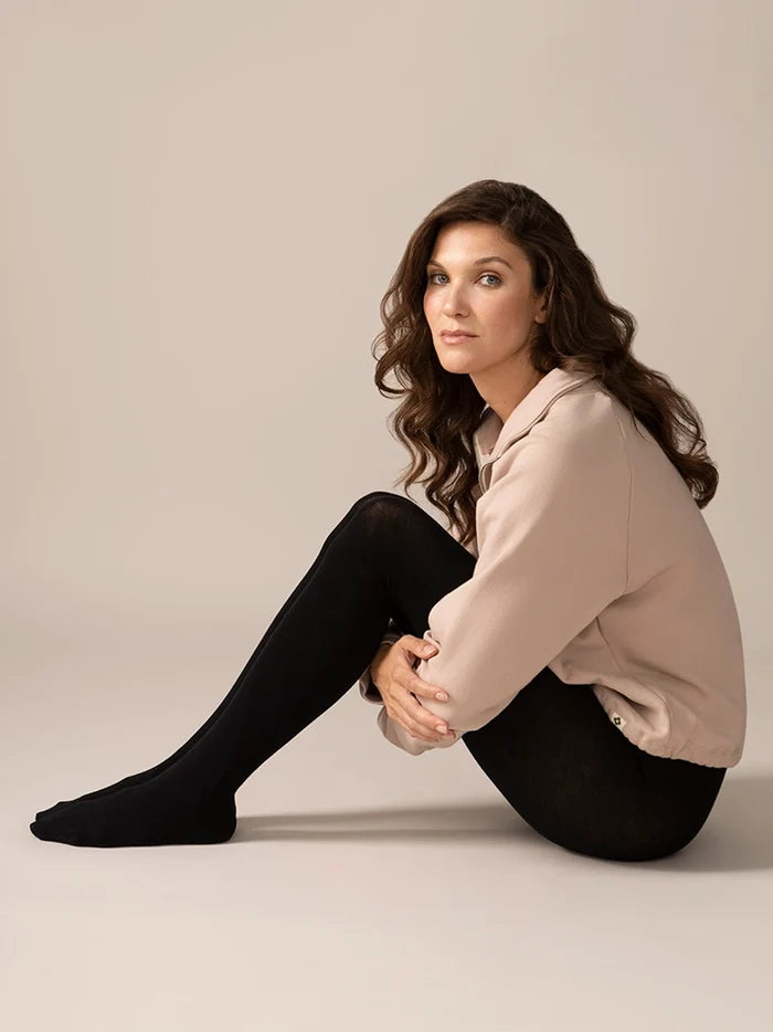 Woman sitting on the floor wearing black tights and a beige jacket against a plain background