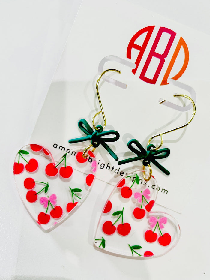 Amanda Bright Designs pair of heart-shaped acrylic dangle earrings with red cherries, green stems, and small pink bows, hanging from gold hooks with dark green ribbon bows.