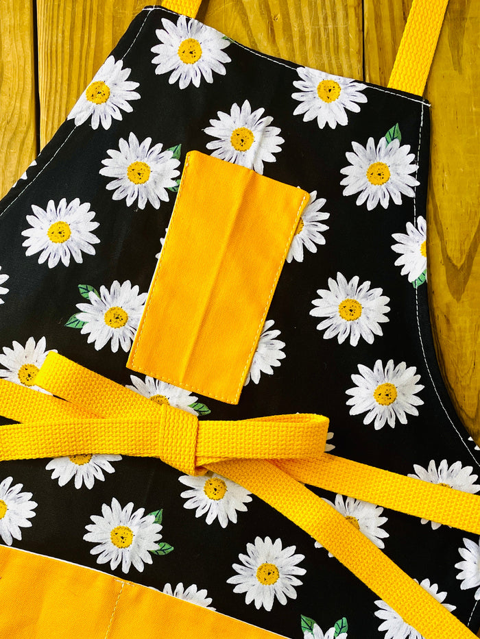 Amanda Bright Designs white daisy with yellow centers pattern apron on black with gold strings and pockets