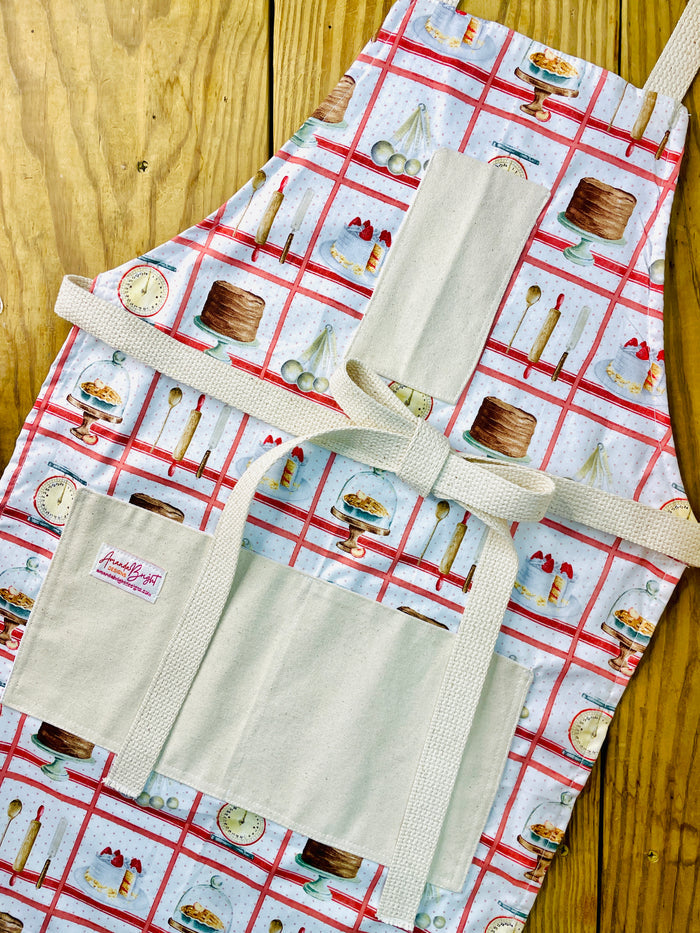 Amanda Bright Designs white baking apron with a red checkered pattern featuring illustrations of baking-related items like a scale, cake, pie, and rolling pin domed cupcakes with canvas pockets and cotton strings