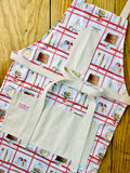 Amanda Bright Designs white baking apron with a red checkered pattern featuring illustrations of baking-related items like a scale, cake, pie, and rolling pin domed cupcakes with canvas pockets and cotton strings