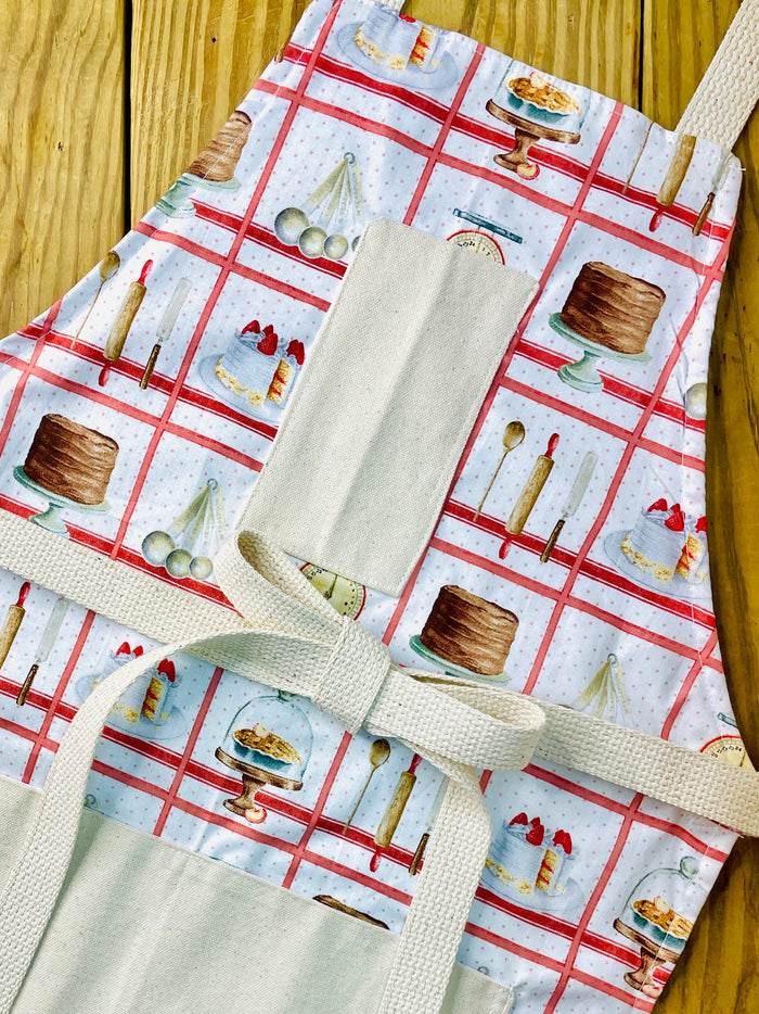 Amanda Bright Designs white baking apron with a red checkered pattern featuring illustrations of baking-related items like a scale, cake, pie, and rolling pin domed cupcakes with canvas pockets and cotton strings