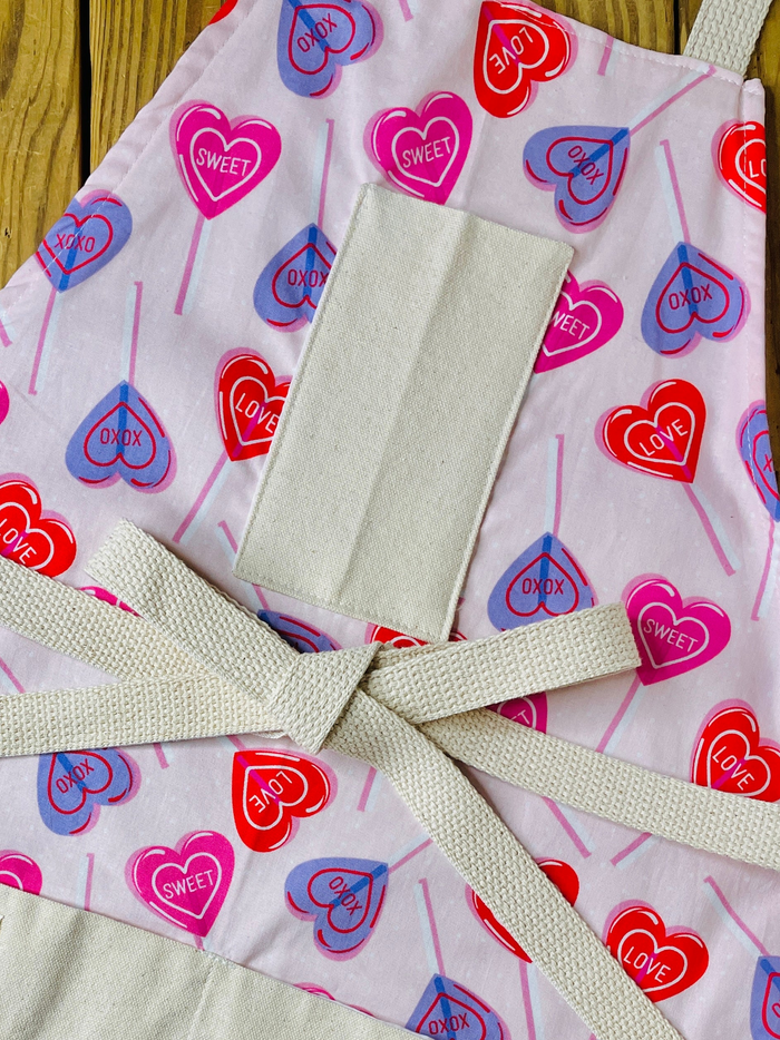 amanda bright designs 100% cotton original design heart lollipop apron Valentine's Day heart pattern, featuring red, pink, and purple heart lollipops with 'CUPID', 'LOVE', and 'SWEET' printed inside