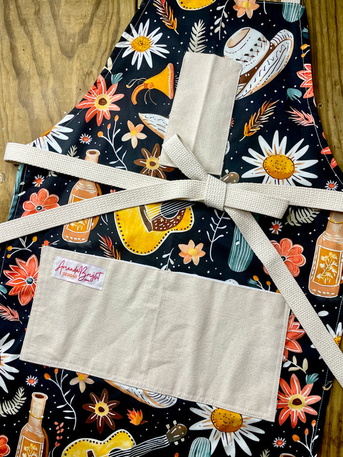 Amanda Bright Designs Desert Cowgirl Apron with colorful western guitar cactus cowboy hat sunflower pattern on black background