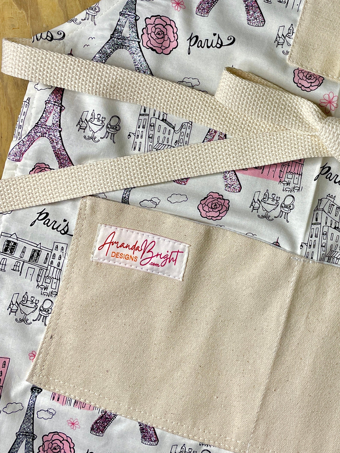 Close-up of apron with a Paris-themed print featuring the Eiffel Tower, buildings, clouds, and pink roses on a cream background.