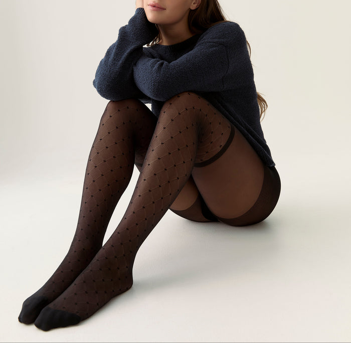 A model sits on a seamless light background wearing sheer black polka-dot tights, a dark navy knit sweater, and black underwear.