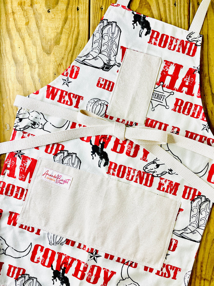 Amanda Bright Designs Cowboy-themed aprons with text and graphics Jane Long Mother of Texas boots, steer, sheriff badge, stars, horseshoes, roping cowboy, horse