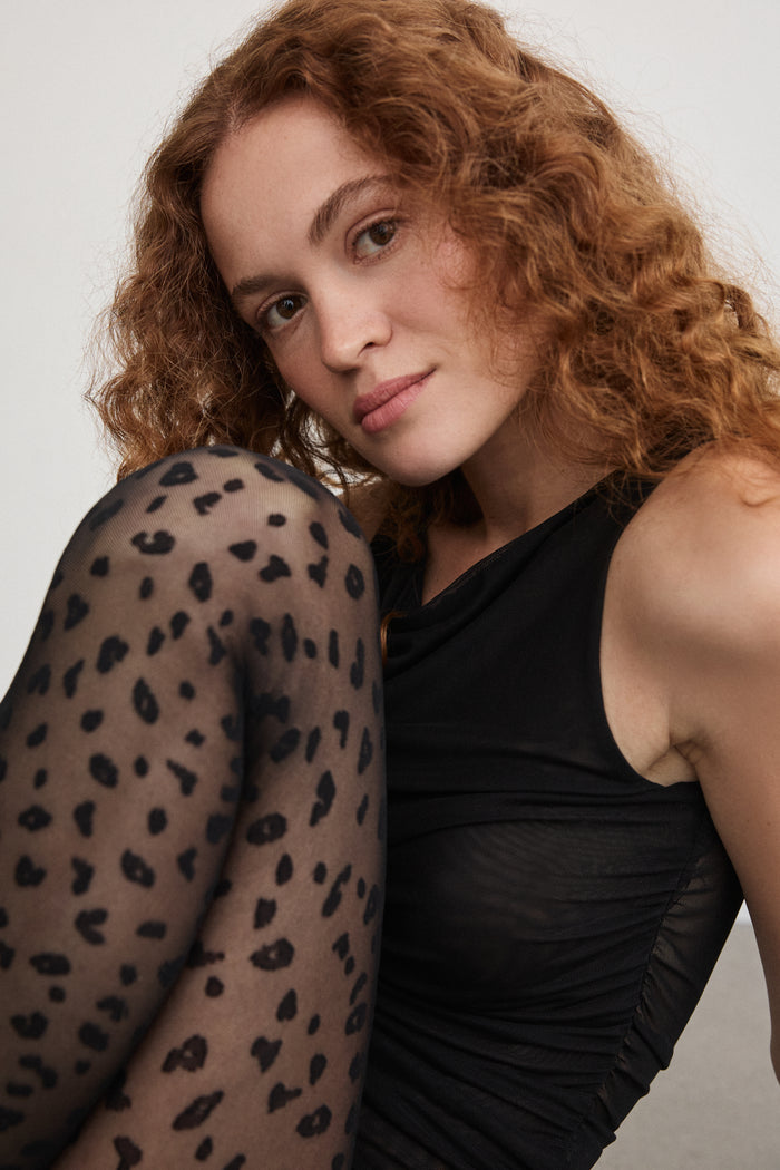 Model wearing Rachel's Mini Leopard Tights in Black with a fitted black top, seated pose showing the leopard print detail.