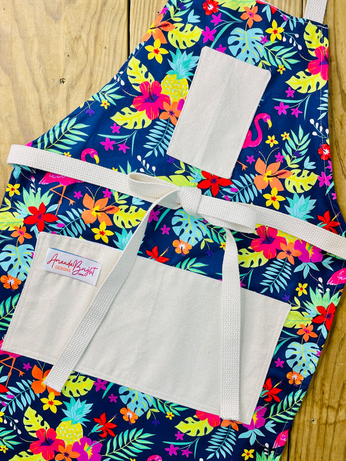 Amanda Bright Designs Nikki multicolored tropical Florida Hawaii floral apron with pink flamingo patterns on a blue background, featuring cotton strings and canvas pockets