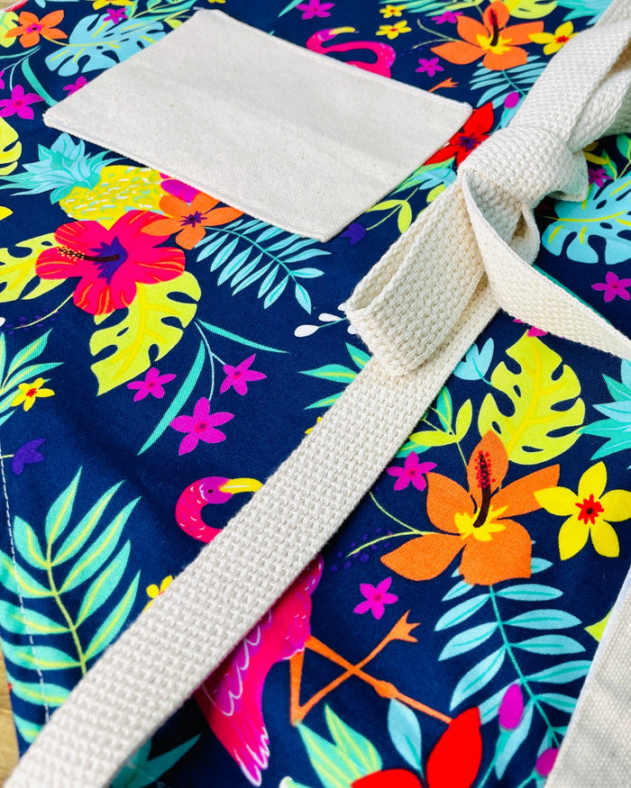 Amanda Bright Designs Nikki multicolored tropical Florida Hawaii floral apron with pink flamingo patterns on a blue background, featuring cotton strings and canvas pockets