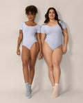 Two women wearing light blue bodysuits against a beige background
