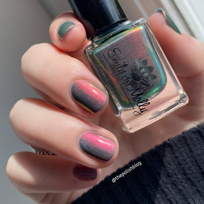 A close-up of a hand with nails painted in a gradient from pink to green, with visible aurora shimmer creating a reflective effect. A bottle of the nail polish is also shown.