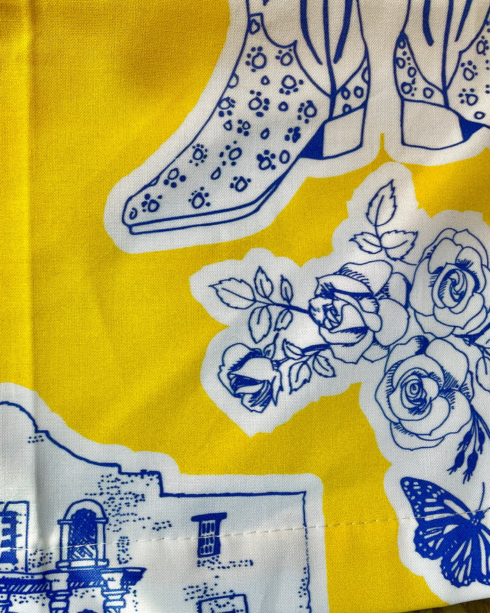 Amanda Bright Designs Texas Toile Apron feaures cowboy boots deer cactus the alamo roses in blue and white on gold background